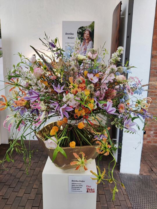 Heimkommen  wenn das bunte Blumenfeld vor Freude tanzt, so heit der Strau von 
<b>Annika Knrr</b>
. Bunt und tanzend, das bringt sie mit einer Vielzahl von Farben und Blumen zum Ausdruck. Ein aufgelockerter Umriss und beschwingt wirkende 
<i>Clematis</i>
, 
<i>Sandersonia</i>
 und 
<i>Gloriosa</i>
 unterstreichen die ungezwungene Ausstrahlung. 