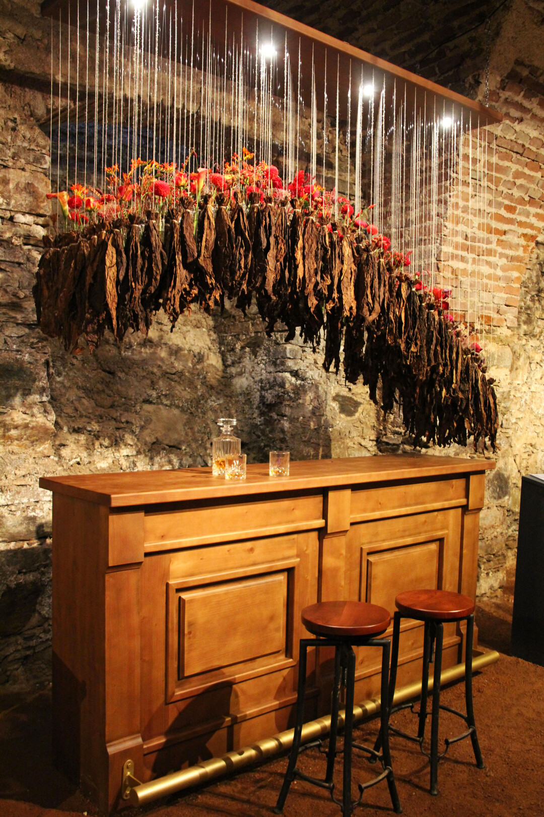 Der Themengestaltung „Ulme“ von
<b>Barbara Schildendorfer</b>
lag ein Gespräch mit der Great Elm im Central Park/New York zugrunde. „In meiner Arbeit setze ich mich mit einer der dunkelsten Phasen der US-amerikanischen Geschichte auseinander: dem Bürgerkrieg zwischen den Nord- und Südstaaten, der vor allem durch die Sklaverei geprägt war.“ Sie schuf ein florales Kunstwerk, das über einer Bar eine mit Blumen gefüllte geschwungene Form von Glasgefäßen und gebündelten Tabakblättern zeigte. Die Gläser enthielten rote Rosen,
<i>Nigella</i>
, Knöterichsorten, Astilben, dunkelrote
<i>Helleborus </i>
u.v.m. Die Auswahl der Farben war dabei entscheidend. Rot steht für den Krieg und das unendlich viele Blut, das vergossen wurde. Die wechselnde Farbqualität beschreibt die unterschiedlichen Phasen des Krieges.